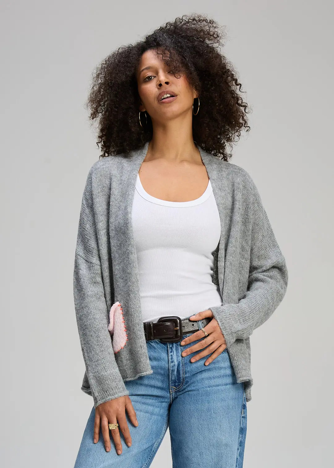 Woman wearing a gray cardigan, white tank top, and blue jeans against a plain background