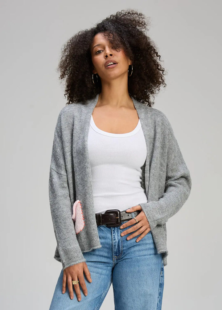 Woman wearing a gray cardigan, white tank top, and blue jeans against a plain background