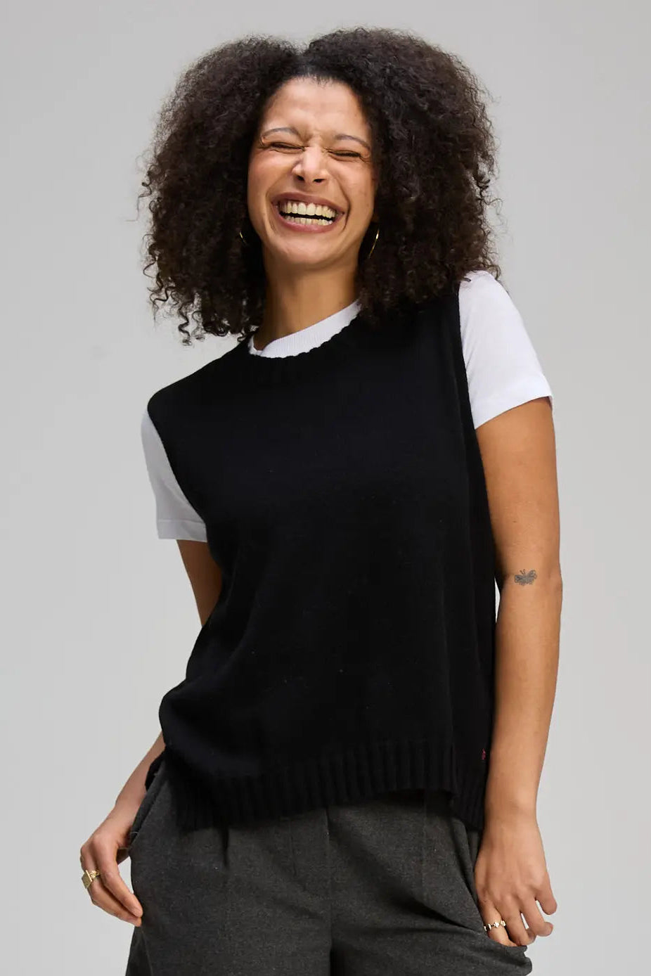 Woman wearing a black sleeveless sweater over a white shirt with gray pants on a plain background. Essential Vest Black
