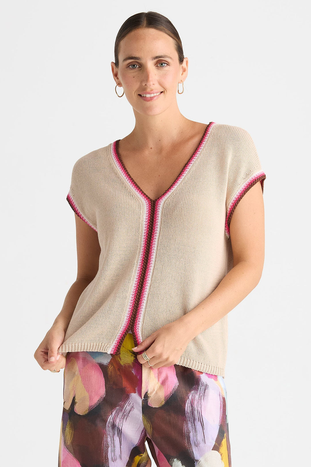 A woman modeling a beige knit V-neck sweater with pink and green striped trim, paired with colorful floral pants, against a plain light gray background.