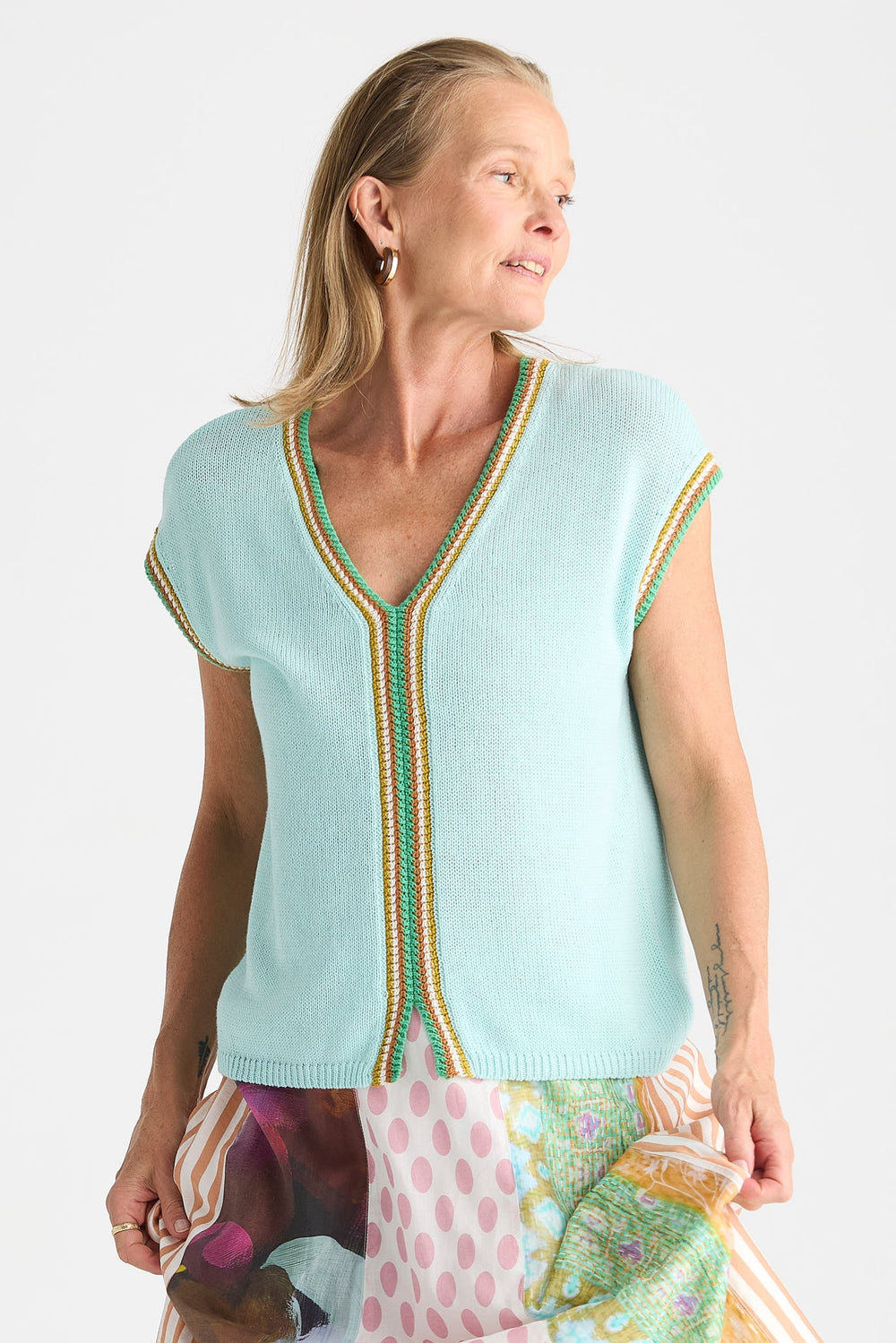 A woman wearing a light aqua knit V-neck sweater vest with green and gold striped trim, paired with a colorful patterned skirt, against a plain white background.