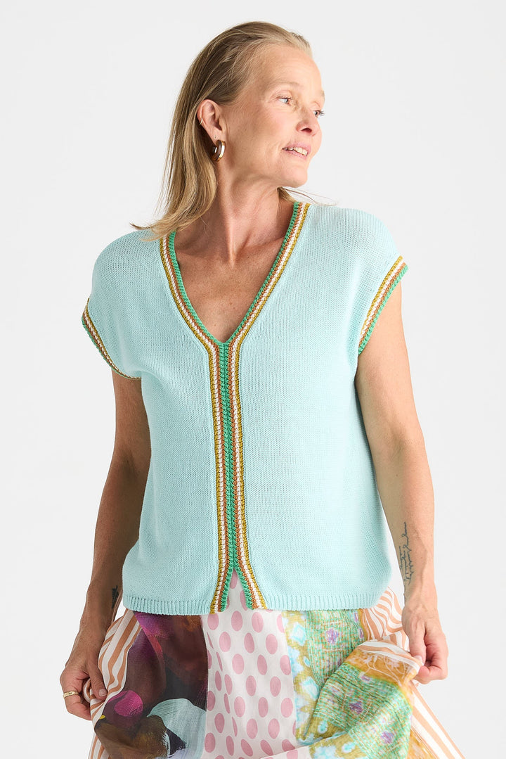 A woman wearing a light aqua knit V-neck sweater vest with green and gold striped trim, paired with a colorful patterned skirt, against a plain white background.