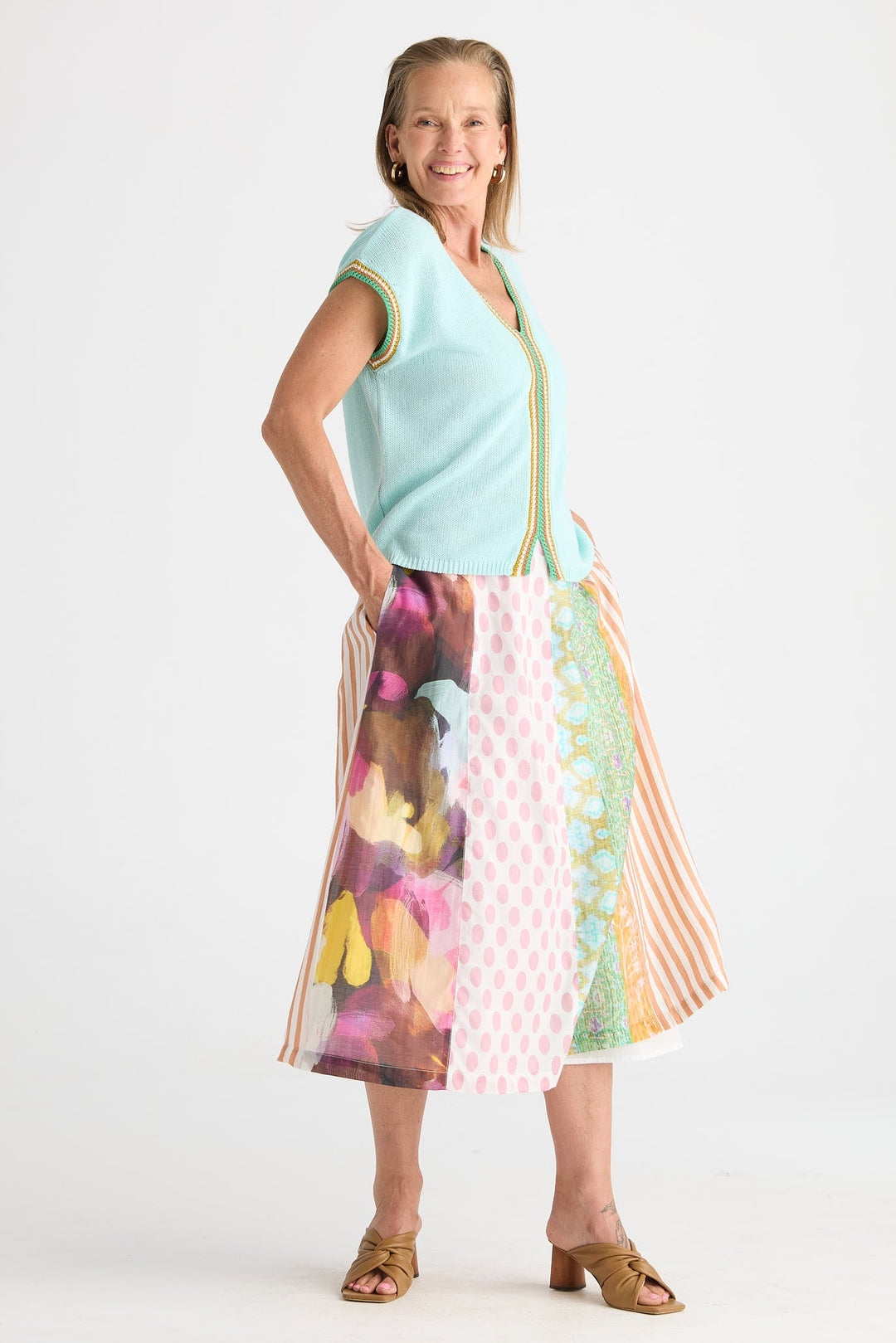 A woman modeling a sleeveless light blue knit vest with gold trim over a colorful midi skirt featuring abstract and striped patterns, paired with brown heeled sandals, against a plain white studio background.