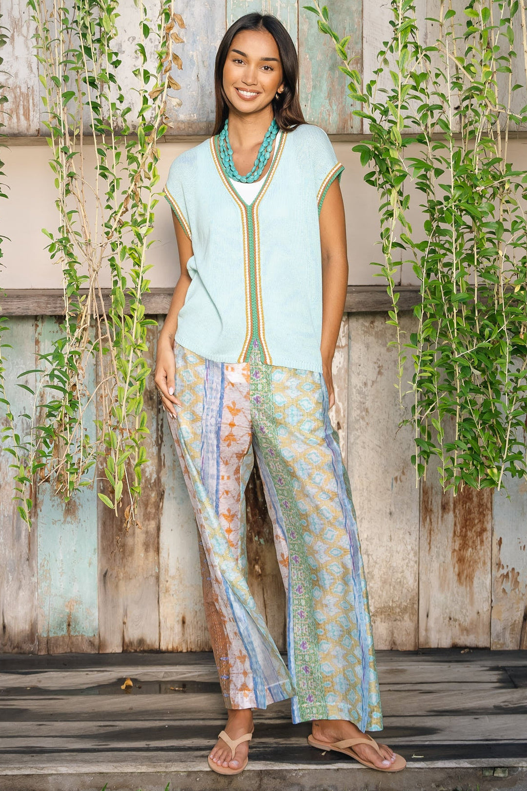 A woman stands outdoors in front of a rustic weathered wooden wall with hanging green vines. She is wearing a sleeveless light blue top with gold trim and matching patterned wide-leg pants in soft pastel tones, along with a turquoise necklace and tan sandals.
