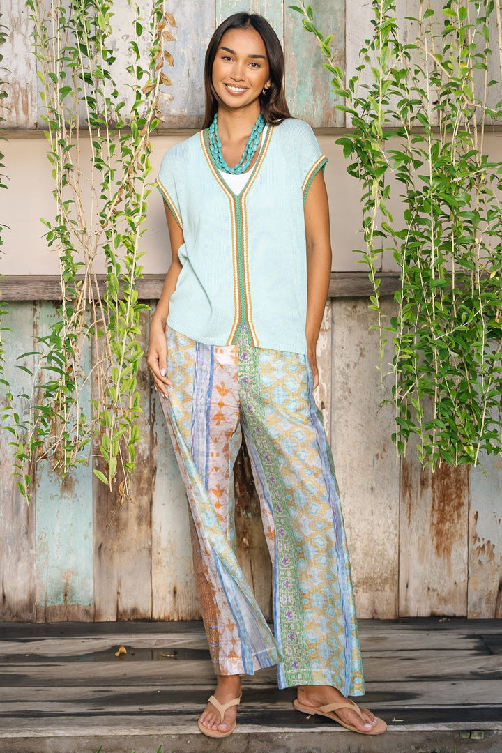 A woman stands outdoors in front of a rustic weathered wooden wall with hanging green vines. She is wearing a sleeveless light blue top with gold trim and matching patterned wide-leg pants in soft pastel tones, along with a turquoise necklace and tan sandals.