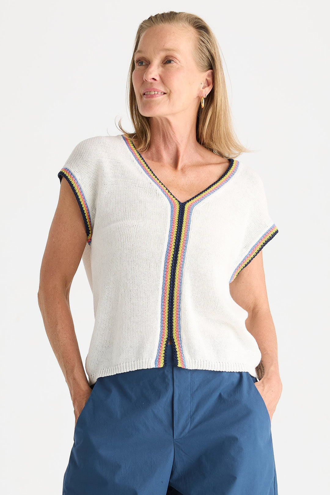 A woman wearing a white knit short-sleeve top with colorful striped trim and blue pants, photographed against a plain light background.