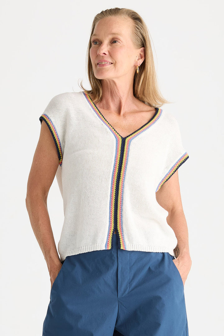 A woman wearing a white knit short-sleeve top with colorful striped trim and blue pants, photographed against a plain light background.