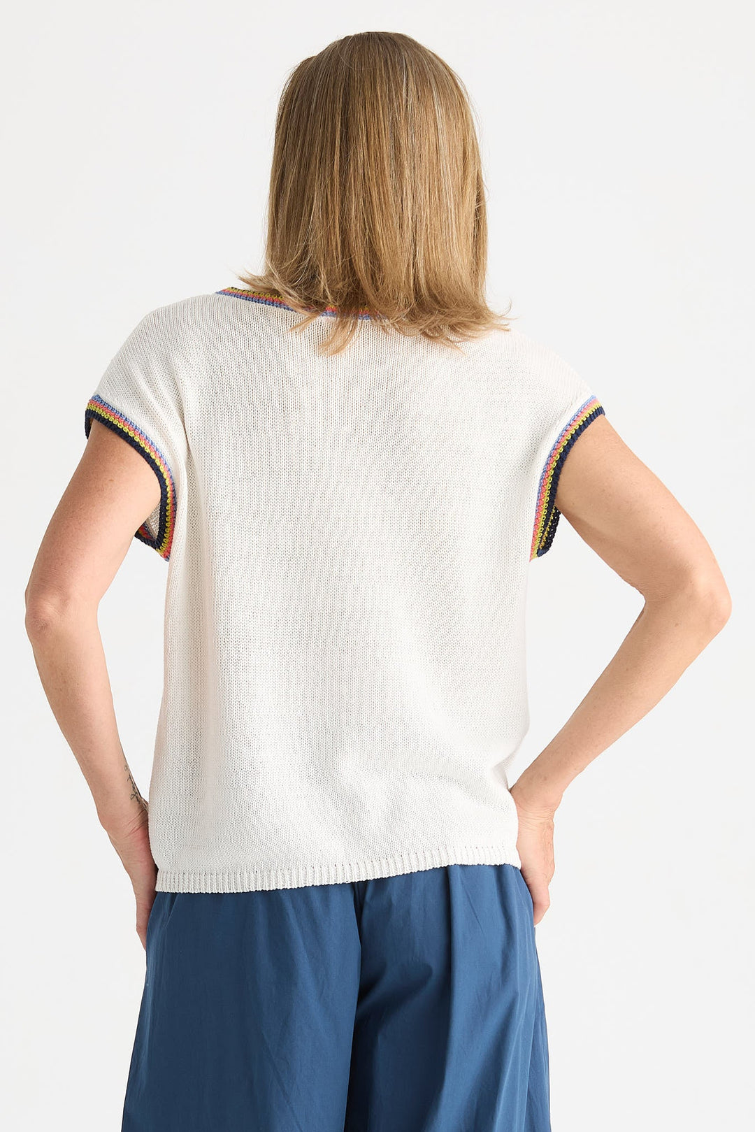 A woman shown from the back wearing a short-sleeve knit top in off-white with striped trim at the sleeve cuffs, paired with blue pants, against a plain light background.
