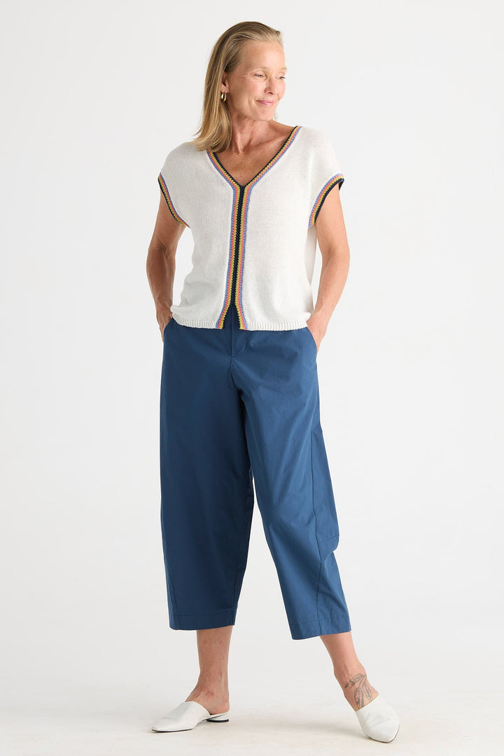 A woman modeling a white short-sleeve top with a thin brown and gold striped trim down the front and along the sleeve cuffs, paired with blue cropped pants and white mule shoes, against a plain white studio background.