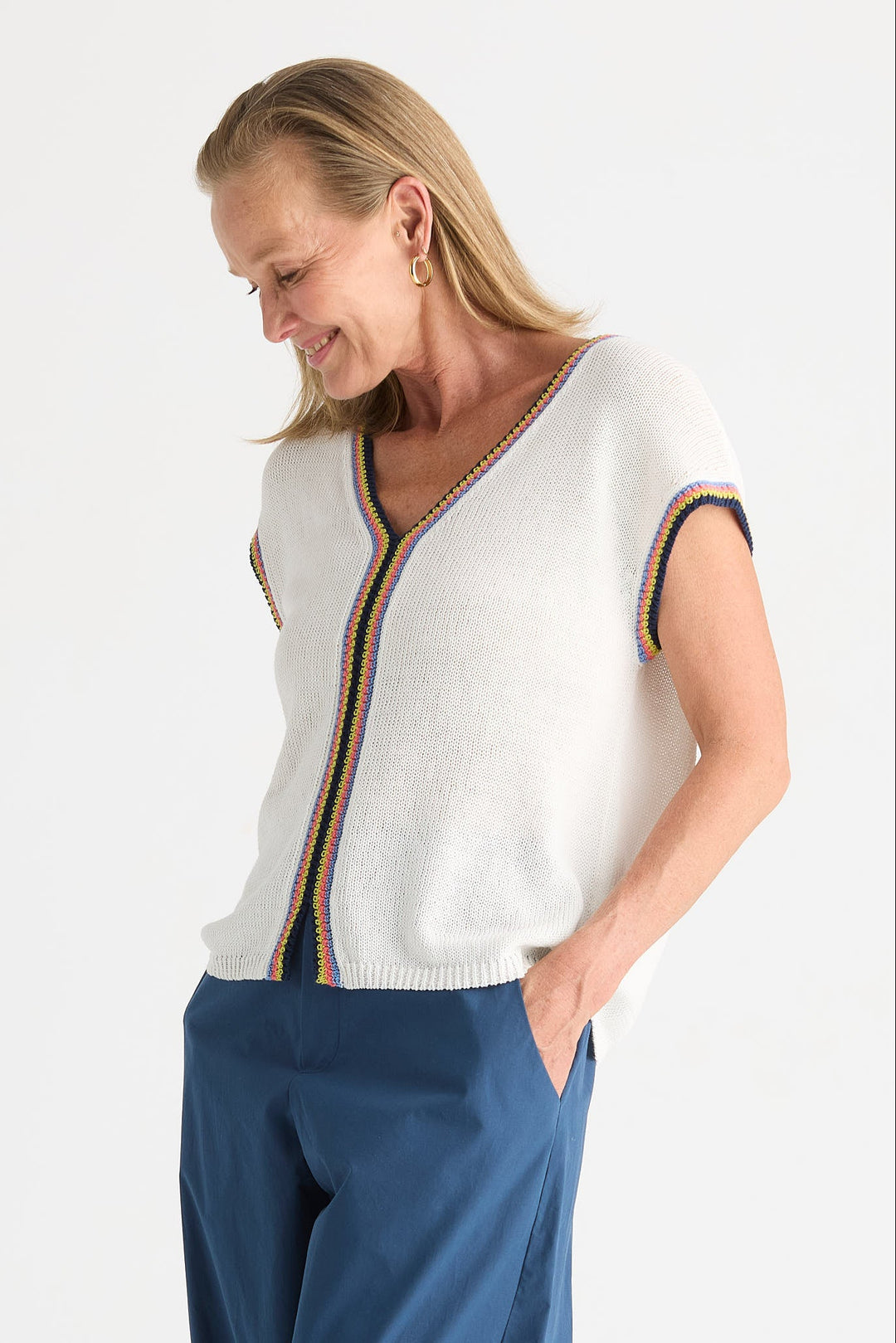 A woman wearing a white knit cardigan with colorful striped trim on the front and sleeve cuffs, paired with navy blue pants, photographed against a plain light background.