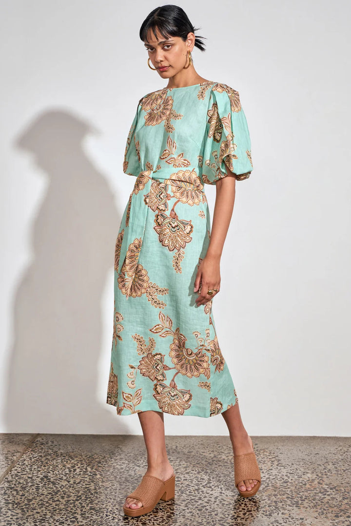 A woman models a mint green midi dress with a brown floral pattern, featuring short puff sleeves and a matching fabric tie belt at the waist, set against a plain white wall.