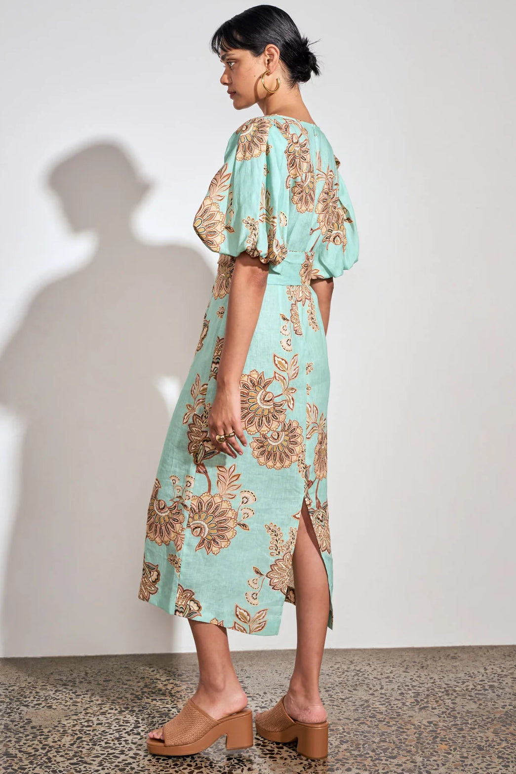 A woman modeling a light aqua midi dress with a brown floral pattern, puff sleeves, and a side slit, standing against a white wall with a shadow.