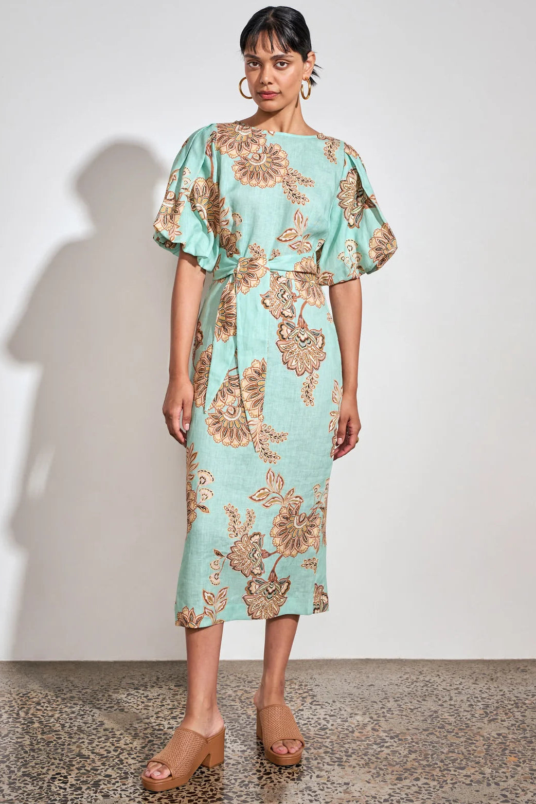 A woman models a light mint green midi dress with a brown floral pattern, featuring puffed short sleeves and a tie at the waist, standing against a plain white wall.