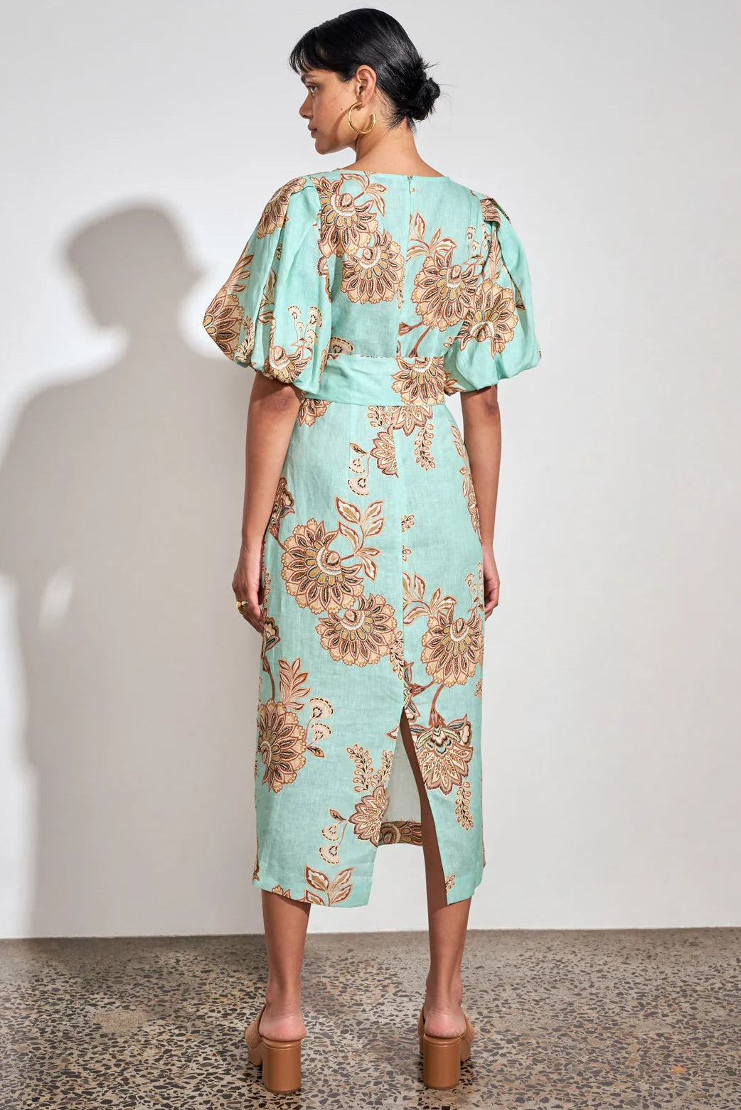 A woman stands with her back to the camera wearing a mint green midi dress with a brown floral pattern and puff sleeves, paired with brown platform sandals, against a white wall.