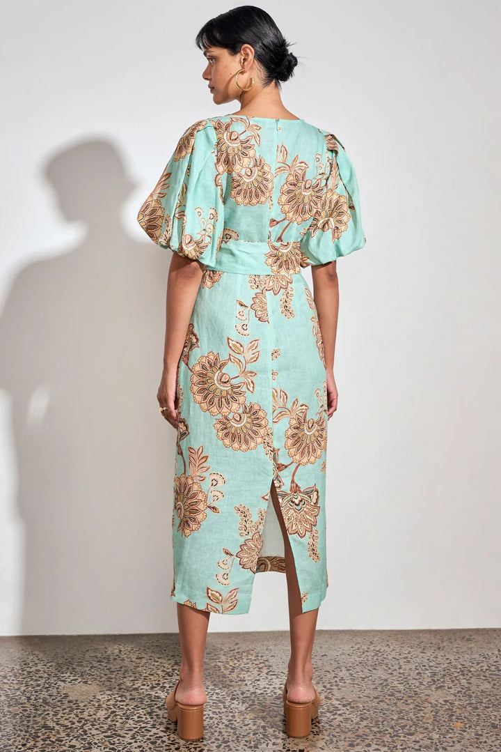 A woman stands with her back to the camera wearing a mint green midi dress with a brown floral pattern and puff sleeves, paired with brown platform sandals, against a white wall.