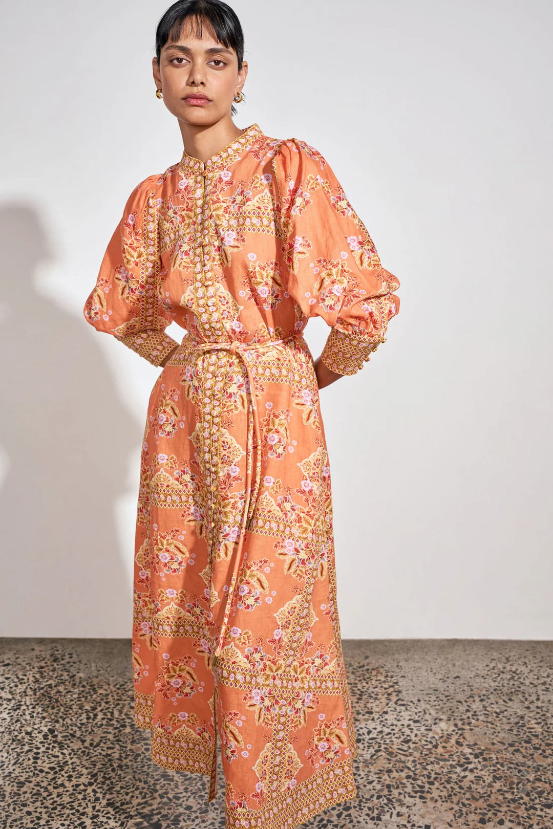 A model wearing a peach-orange maxi dress with a detailed gold, pink, and green floral and paisley print, standing against a plain off-white wall.