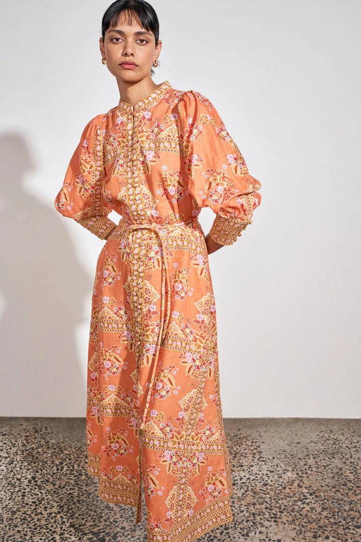 A model wearing a peach-orange maxi dress with a detailed gold, pink, and green floral and paisley print, standing against a plain off-white wall.