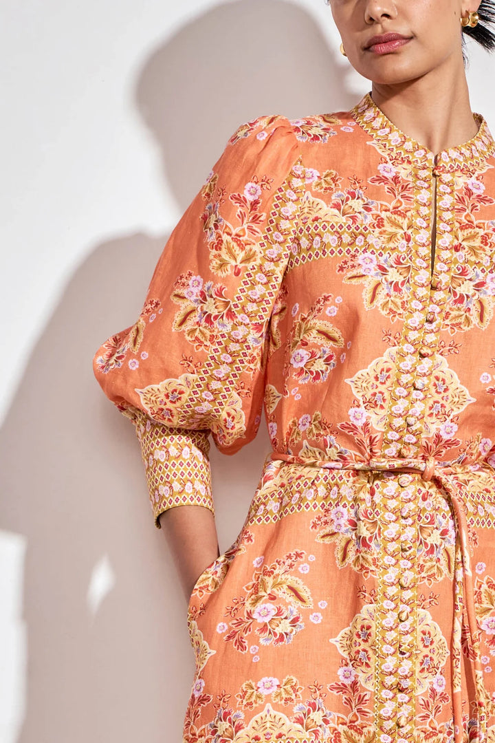 A close-up of a model wearing a peach-orange dress with a detailed floral and geometric print in pink, green, and gold, featuring balloon sleeves and a tie waist.