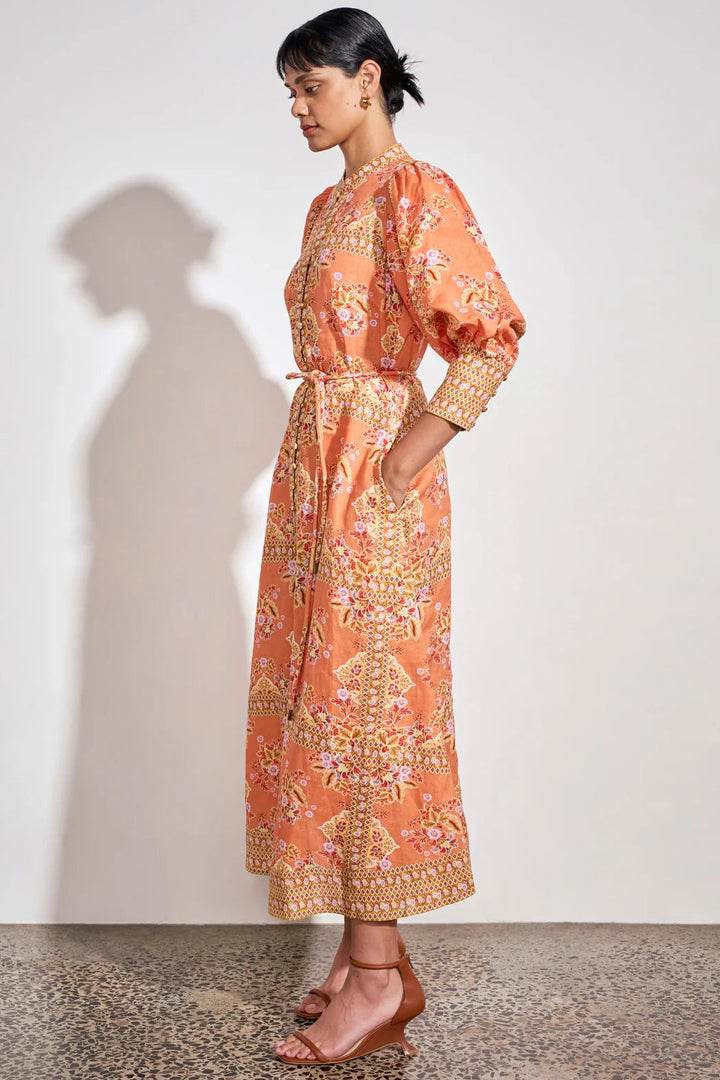 A woman models a peach-orange maxi dress with a detailed gold and pink floral pattern, featuring puff sleeves and a matching waist tie, set against a plain white wall with a soft shadow.