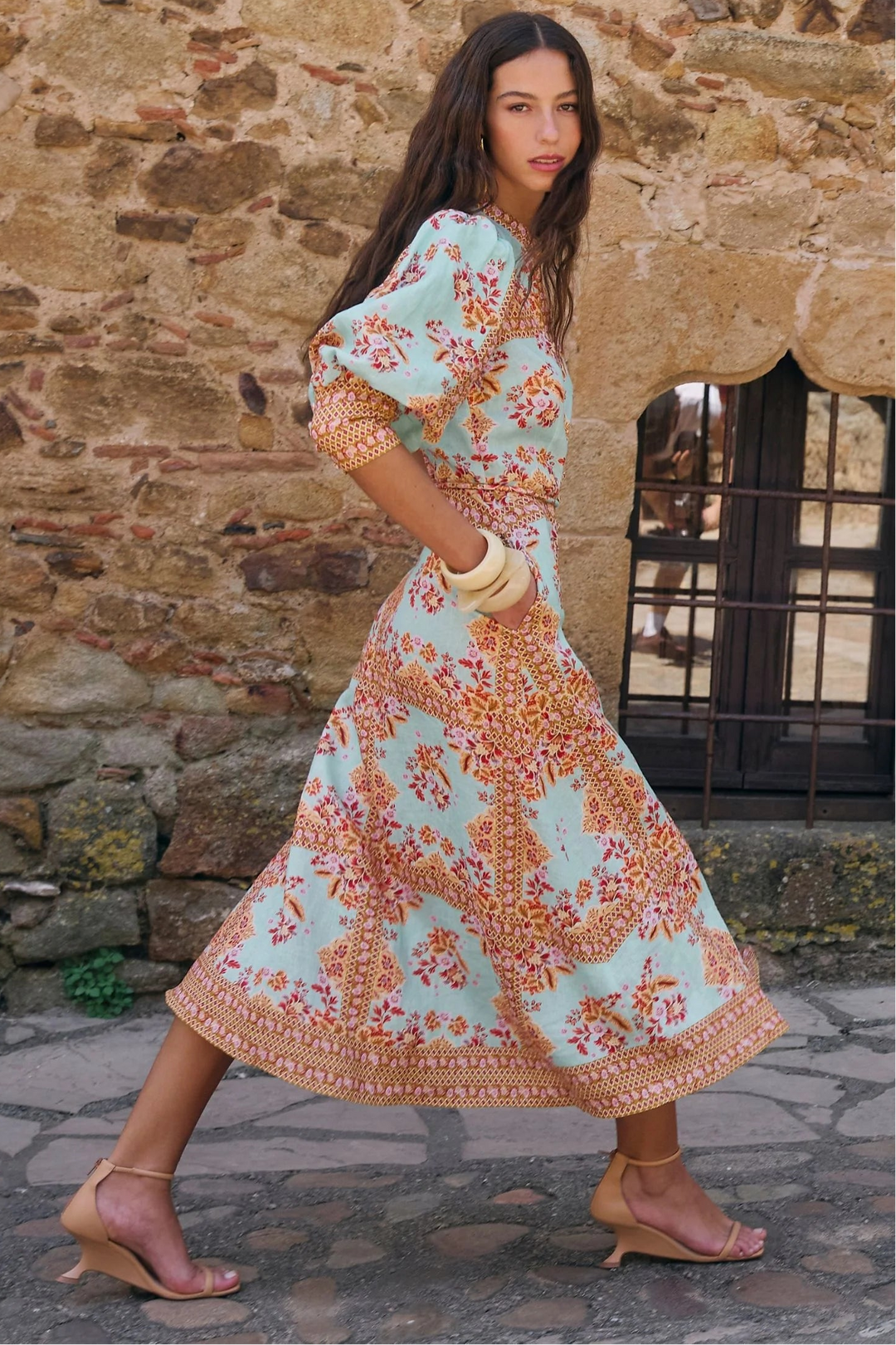 A woman models a light blue and orange floral print maxi dress with puff sleeves, standing in front of a rustic stone wall.