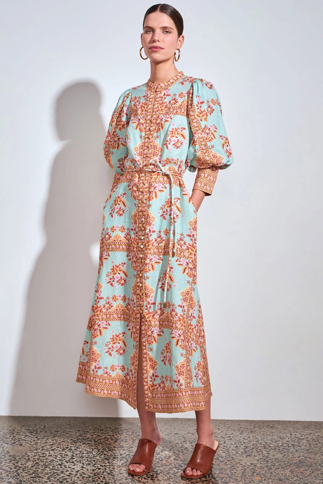A woman models a long-sleeved midi dress with a light blue-green base and a rust-orange floral pattern, featuring a tie waist and puff sleeves, set against a plain light gray wall.