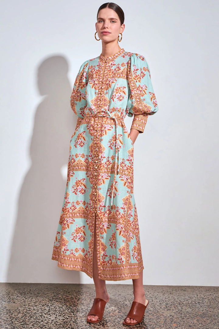 A woman models a long-sleeved midi dress with a light blue-green base and a rust-orange floral pattern, featuring a tie waist and puff sleeves, set against a plain light gray wall.