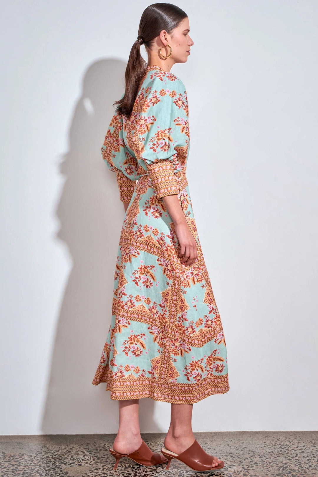 A woman stands in profile wearing a light aqua midi dress with an orange and beige floral pattern, paired with brown leather slingback heels, against a plain white wall.