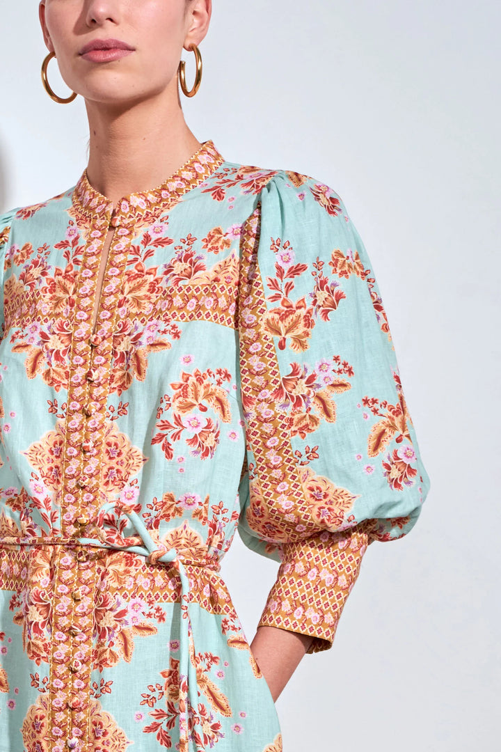 A close-up of a model wearing a light blue-green dress with a vibrant orange, pink, and gold floral pattern, featuring puff sleeves and a matching waist tie.
