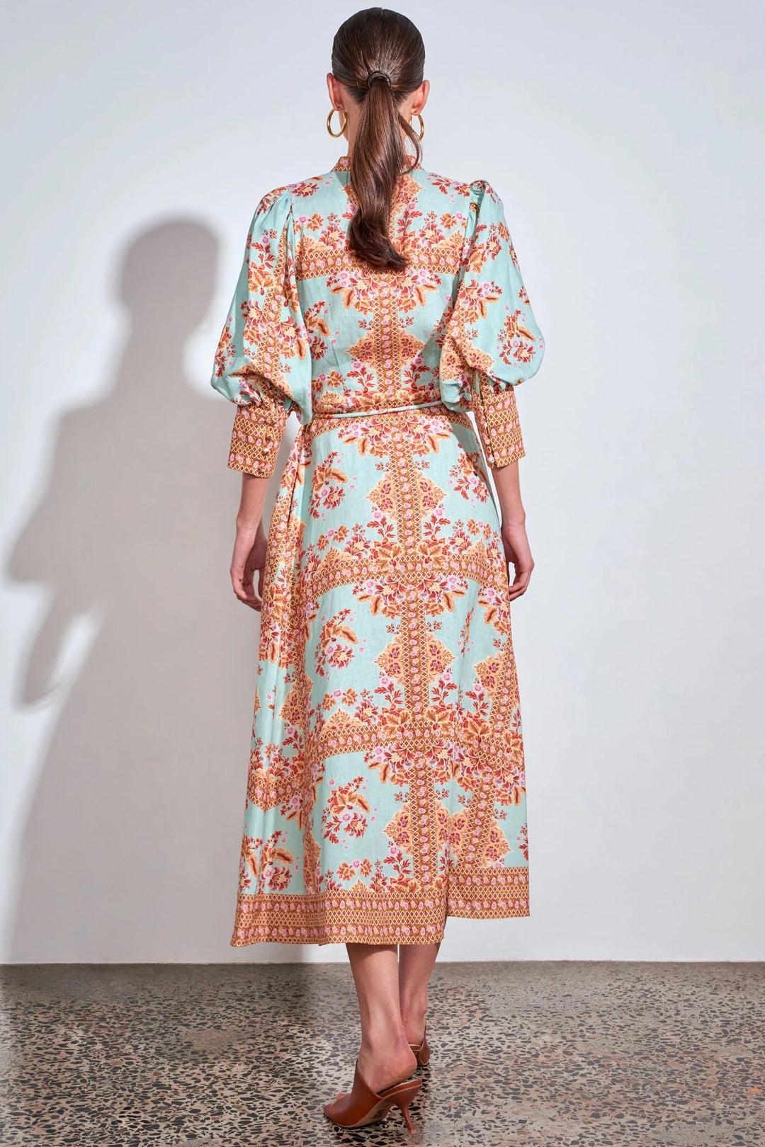 A woman viewed from behind wearing a long-sleeved midi dress with a light blue and orange floral pattern, standing against a plain white wall.