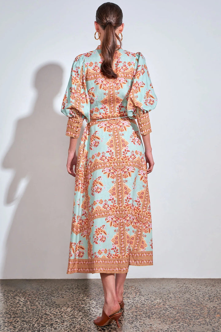 A woman viewed from behind wearing a long-sleeved midi dress with a light blue and orange floral pattern, standing against a plain white wall.