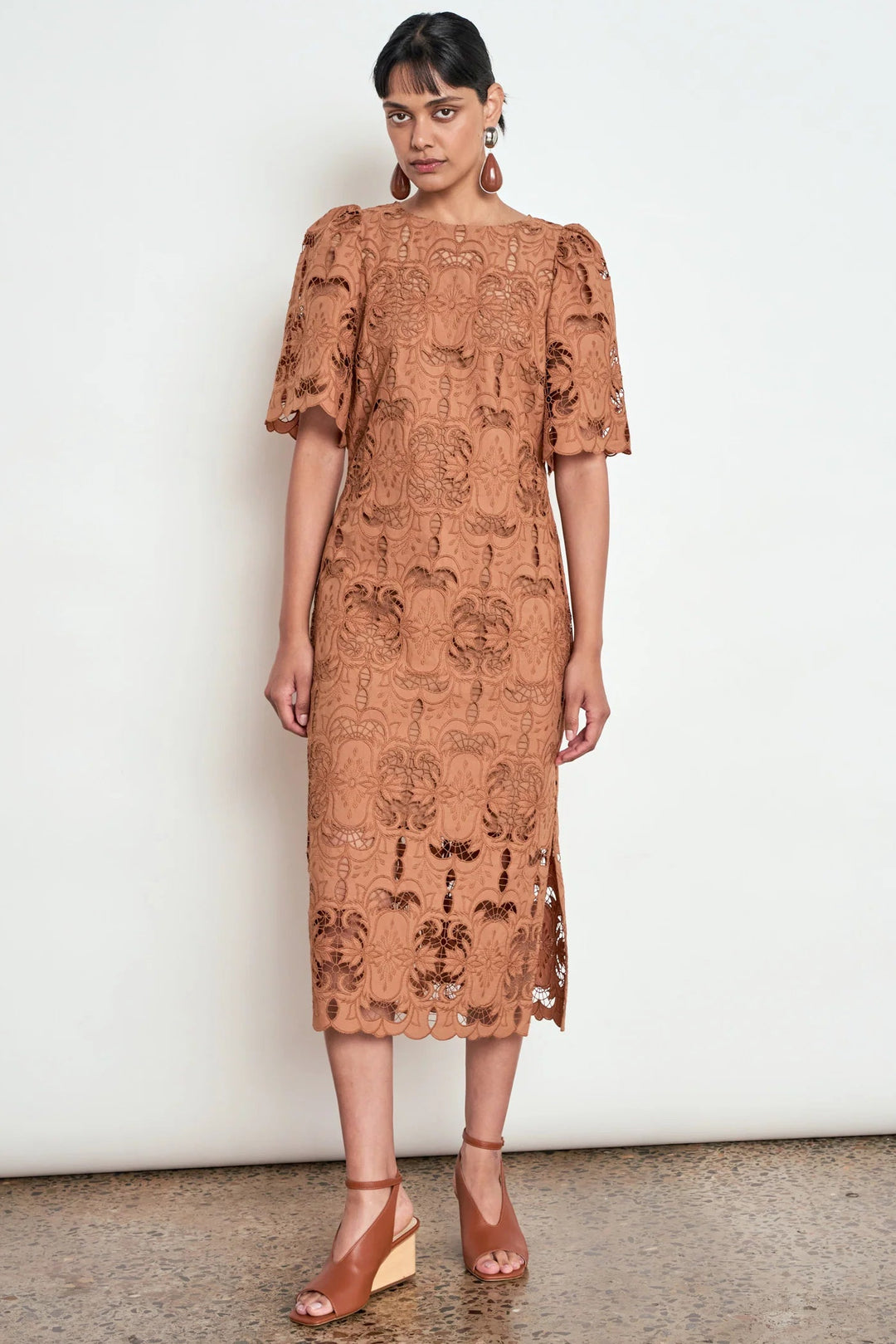 A woman stands against a plain off-white wall wearing a rust-brown embroidered midi dress with puff sleeves and tan wedge sandals.