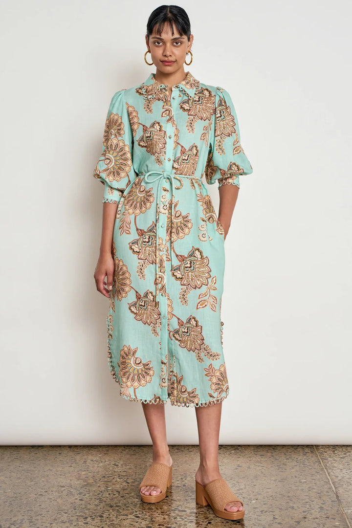 A woman stands against a plain off-white wall wearing a light aqua midi shirt dress with a brown and beige floral paisley pattern, paired with tan wedge sandals.
