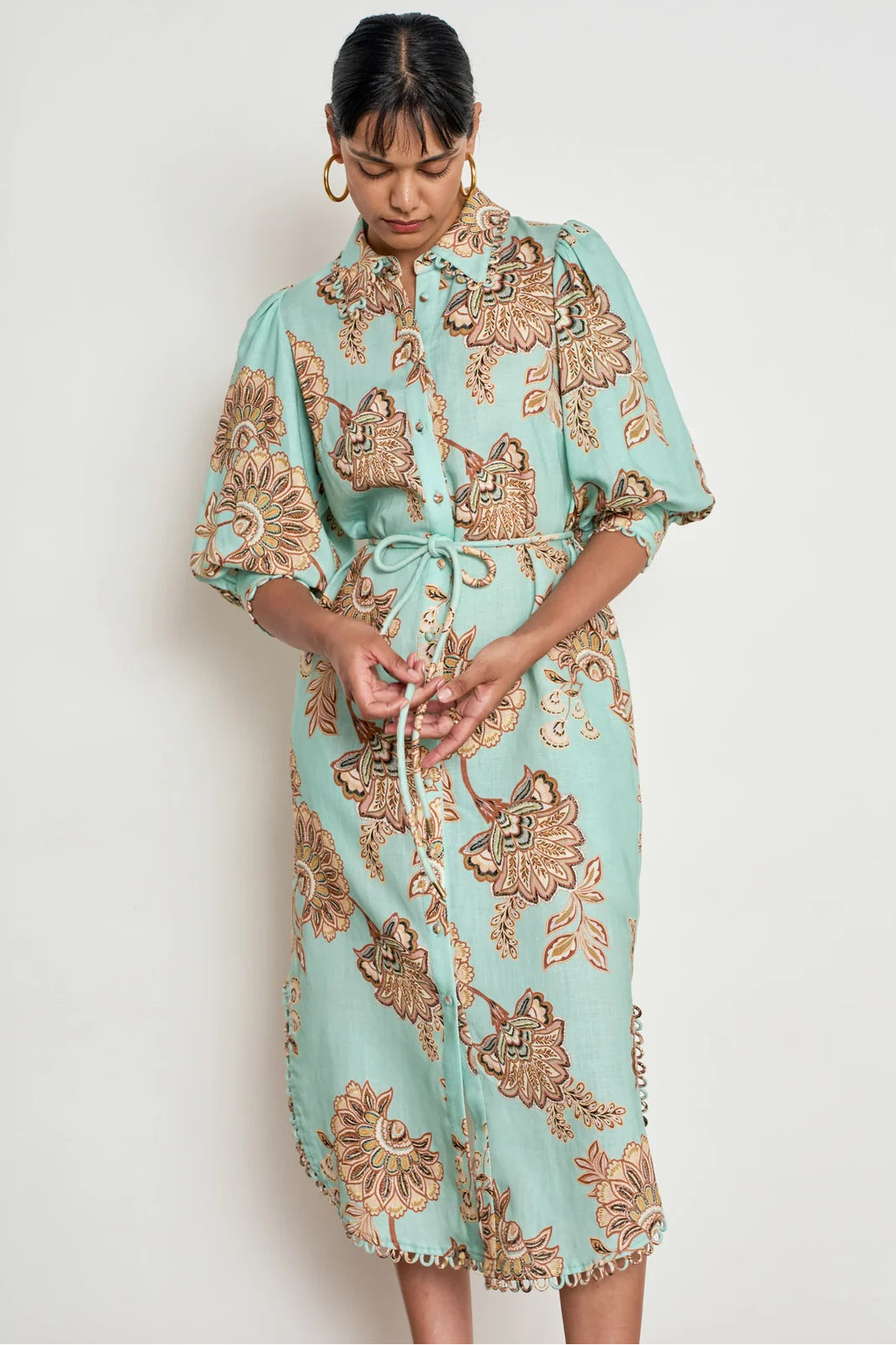 A woman wearing a mint green and brown floral print shirt dress with a tie waist, standing against a plain off-white background.