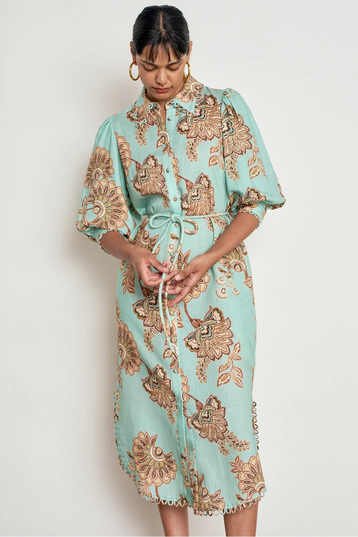 A woman wearing a mint green and brown floral print shirt dress with a tie waist, standing against a plain off-white background.