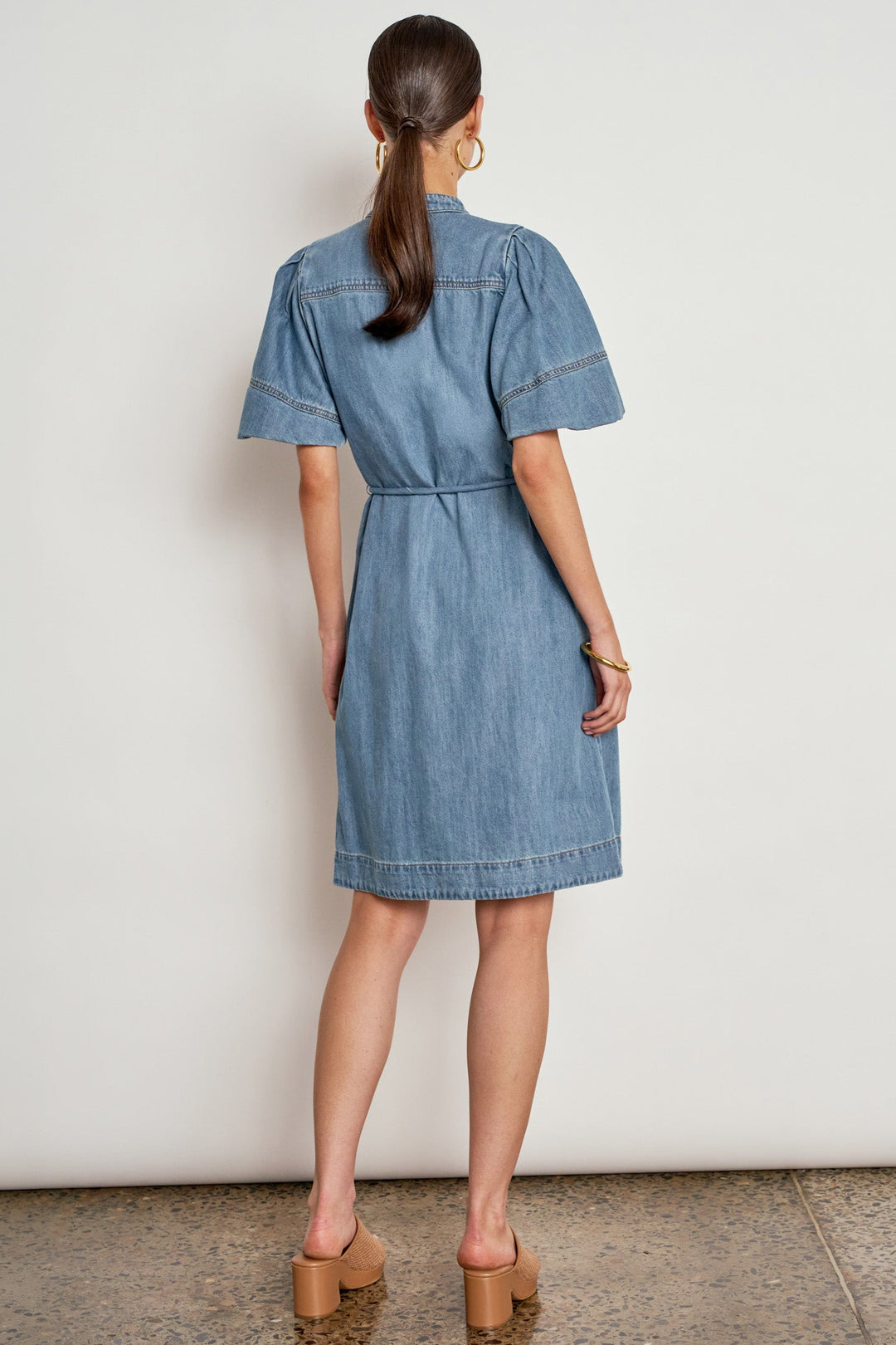 A woman viewed from the back wearing a light blue denim shirt dress with short sleeves, a buttoned collar, and a tie waist, paired with tan block-heel mules against a plain off-white wall.