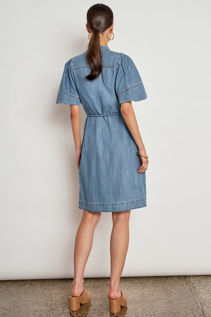 A woman viewed from the back wearing a light blue denim shirt dress with short sleeves, a buttoned collar, and a tie waist, paired with tan block-heel mules against a plain off-white wall.