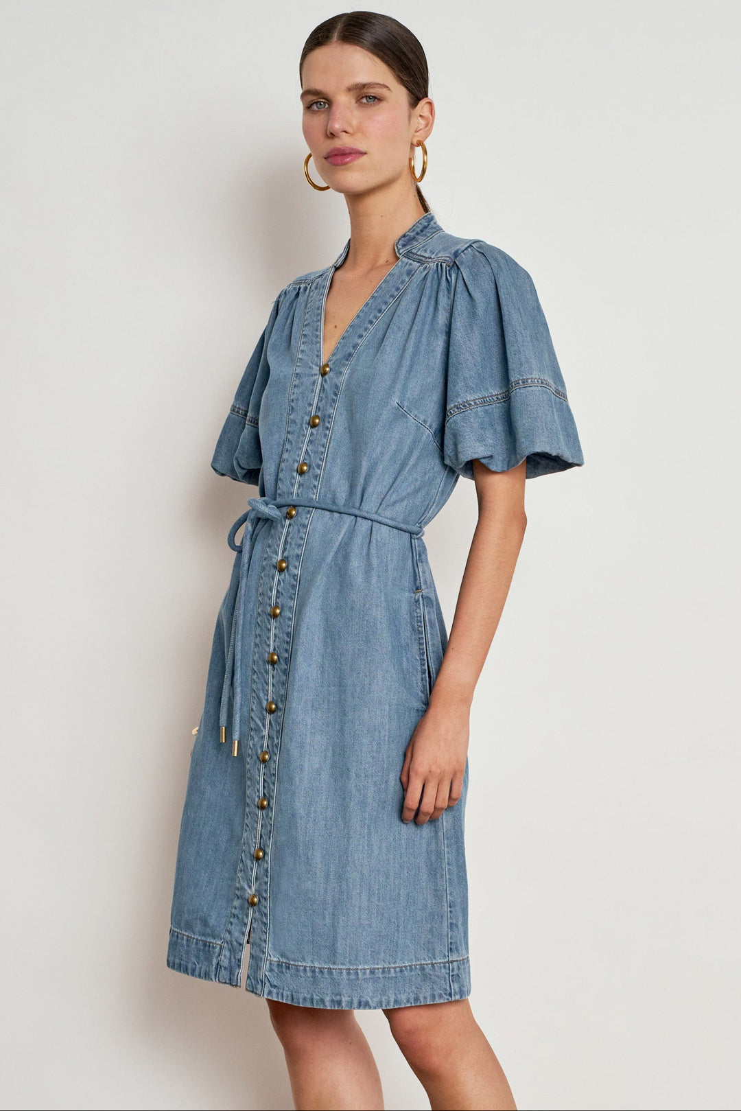 A woman modeling a light blue denim dress with puff sleeves, a button-front bodice, and a matching tie belt, photographed against a plain light gray studio background.