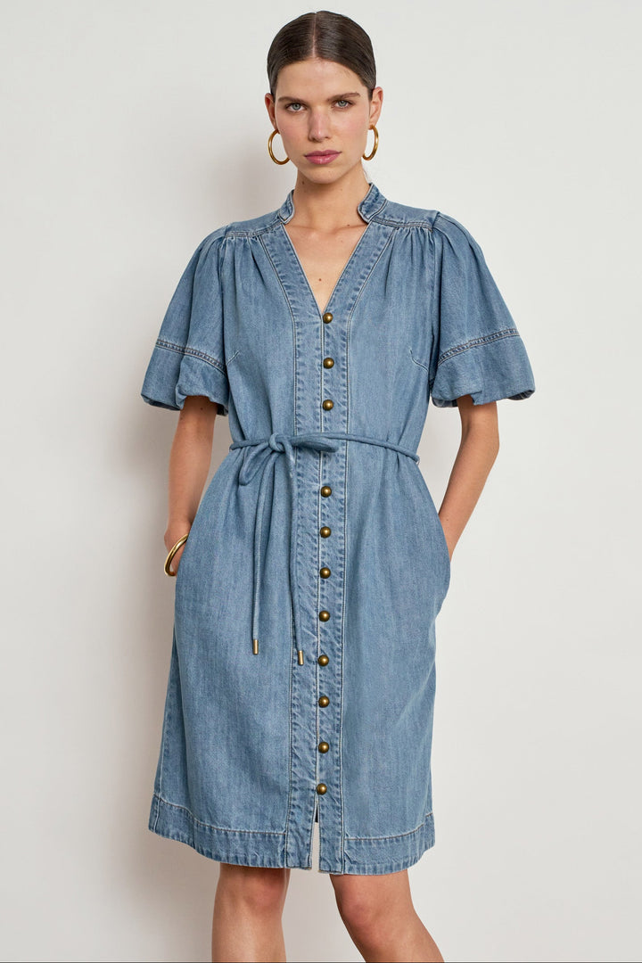A woman modeling a light blue denim shirt dress with short puff sleeves, a V-neckline, button-front closure, and a self-tie waist belt, photographed against a plain light gray studio background.