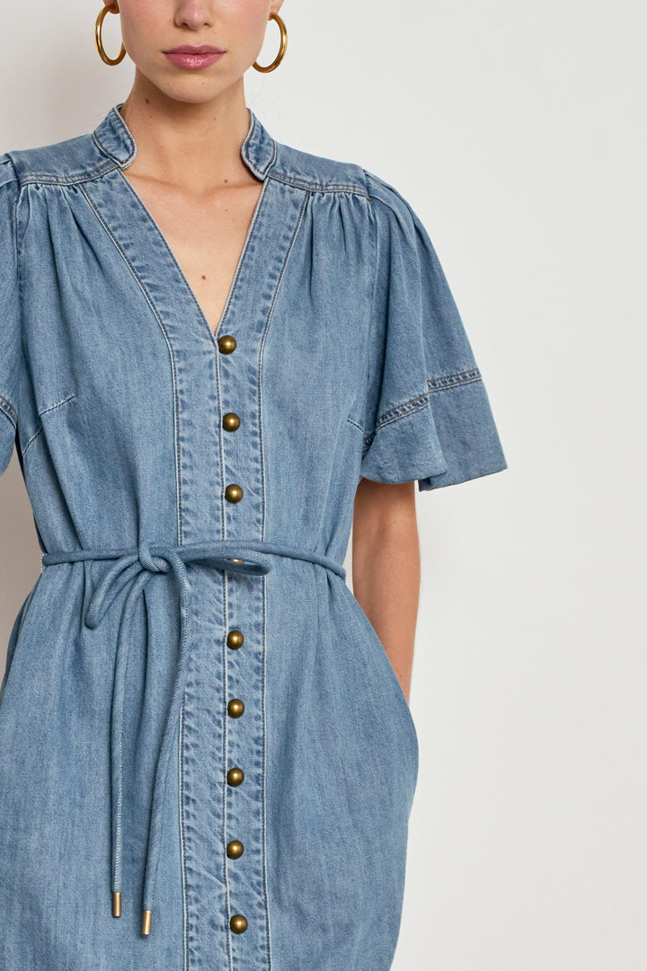 A close-up of a model wearing a light blue denim dress with a V-neck, button-down front, puff sleeves, and a matching tie belt at the waist.