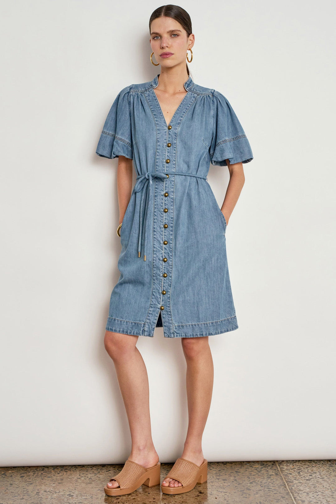 A woman models a light blue denim midi dress with short puff sleeves, a button-front closure, and a self-tie waist belt, standing against a plain off-white wall.