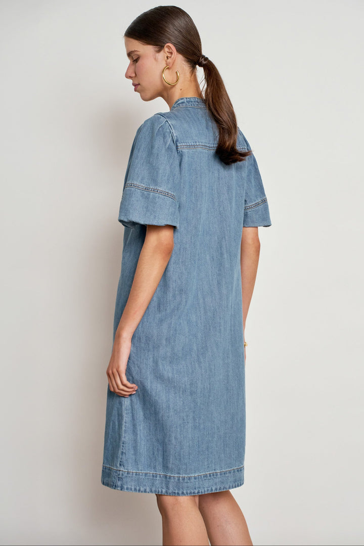 A woman wearing a light blue denim shirt dress, shown from a side-back angle against a plain light gray studio background.