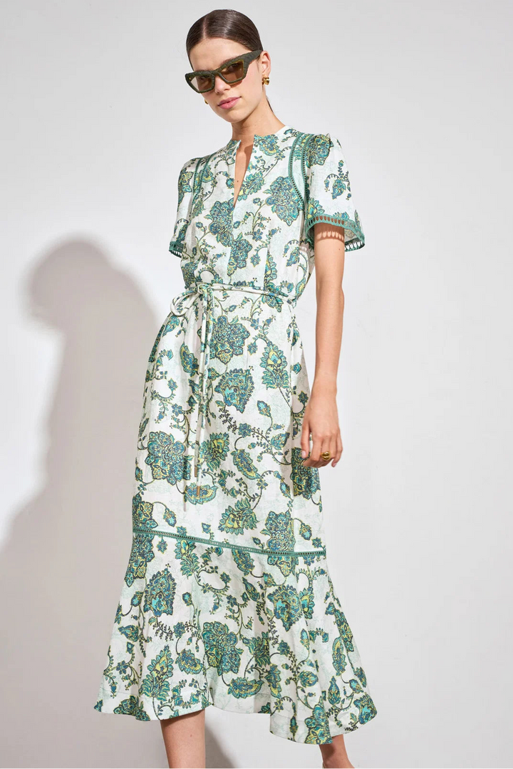 A woman modeling a short-sleeve midi dress with a green and teal floral print on a white background, styled with dark sunglasses against a light gray studio background.