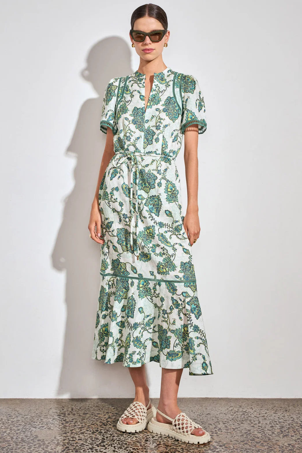 A model wearing a short-sleeved midi dress with a green and white botanical pattern, paired with beige woven sandals and sunglasses, standing against a plain white wall.