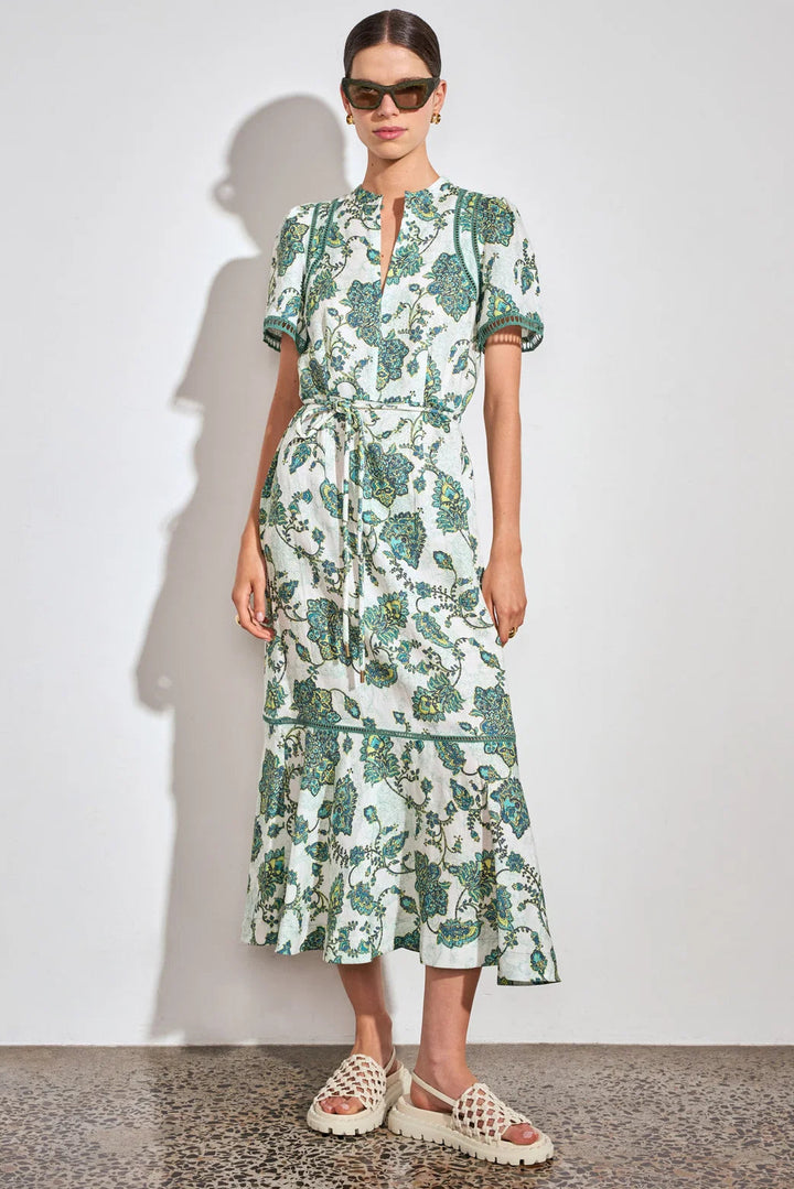 A model wearing a short-sleeved midi dress with a green and white botanical pattern, paired with beige woven sandals and sunglasses, standing against a plain white wall.