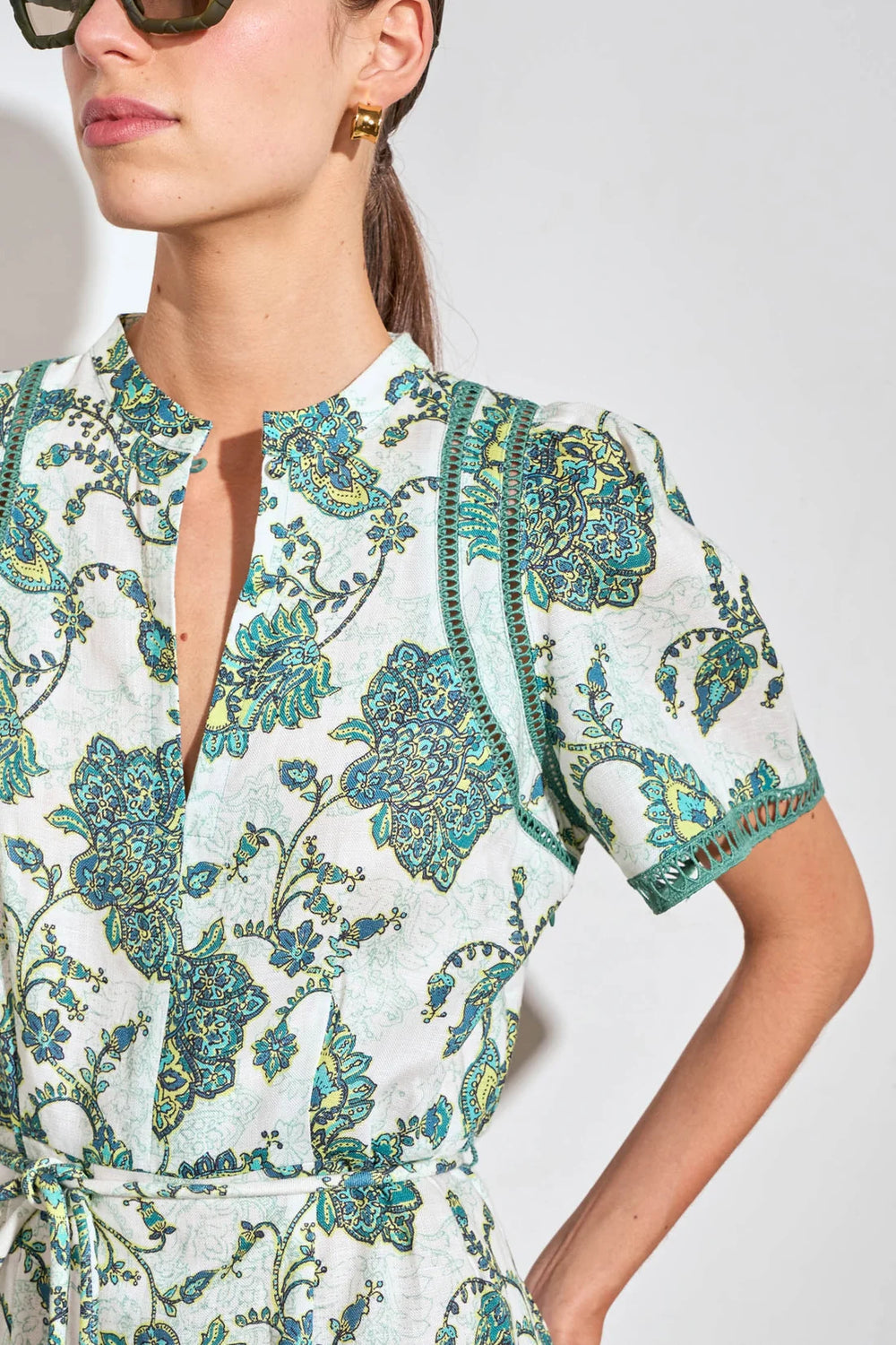 A close-up of a woman wearing a short-sleeve wrap dress with a white base and a teal and green floral paisley pattern. The dress features a split neckline with a keyhole cutout and a matching fabric tie belt at the waist.