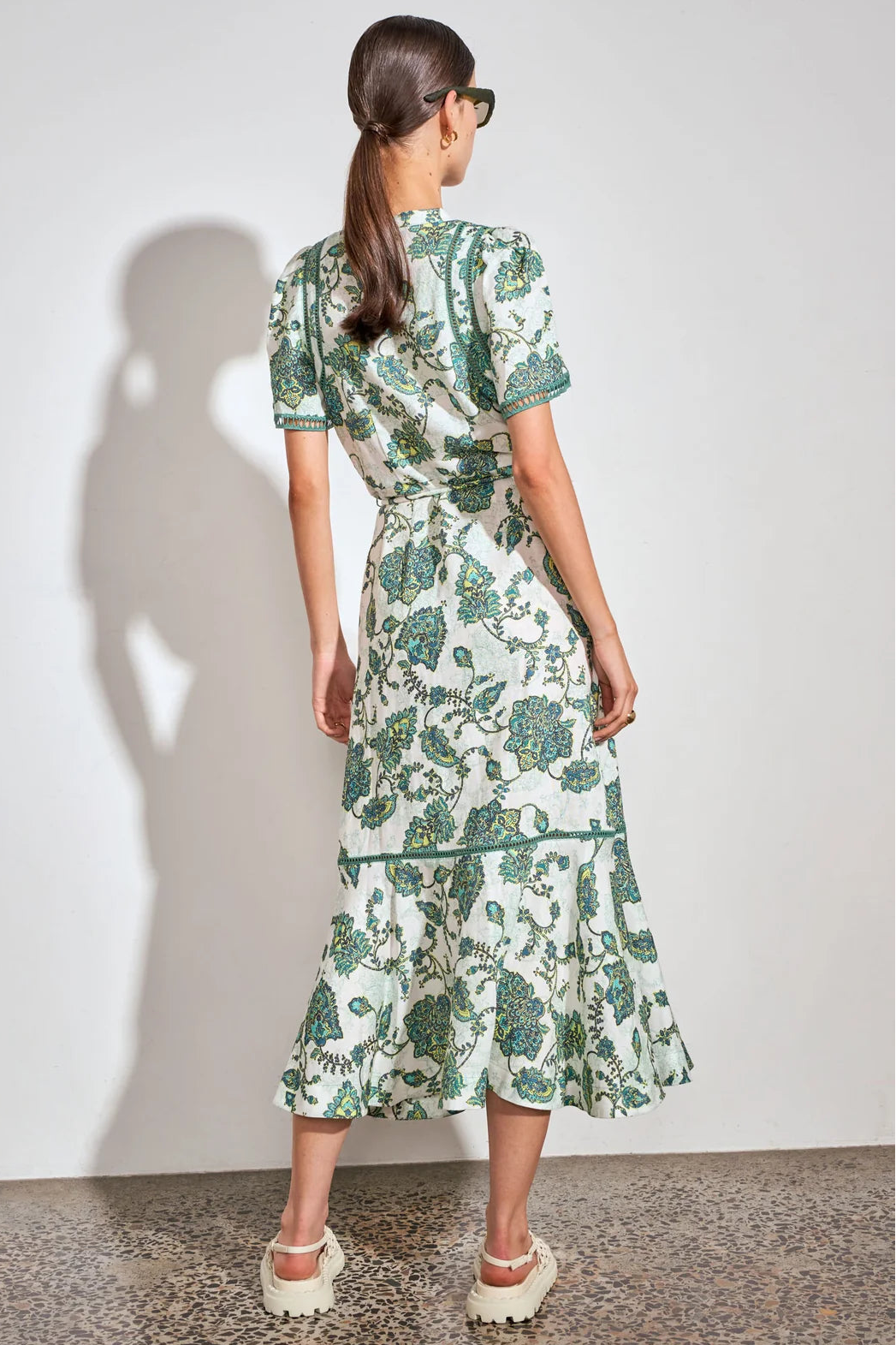 A woman stands with her back to the camera wearing a short-sleeve midi dress with a green botanical print on a white background. She is wearing white platform sandals and standing against a plain white wall with a soft shadow cast on the left.