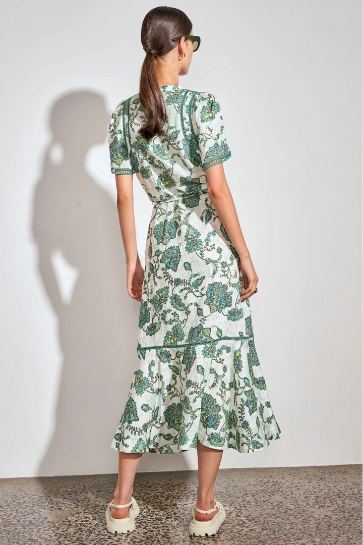 A woman stands with her back to the camera wearing a short-sleeve midi dress with a green botanical print on a white background. She is wearing white platform sandals and standing against a plain white wall with a soft shadow cast on the left.