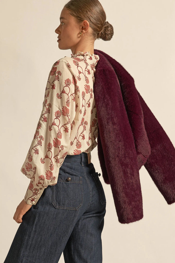 Woman wearing a floral blouse and dark pants with a maroon coat over her shoulder on a beige background