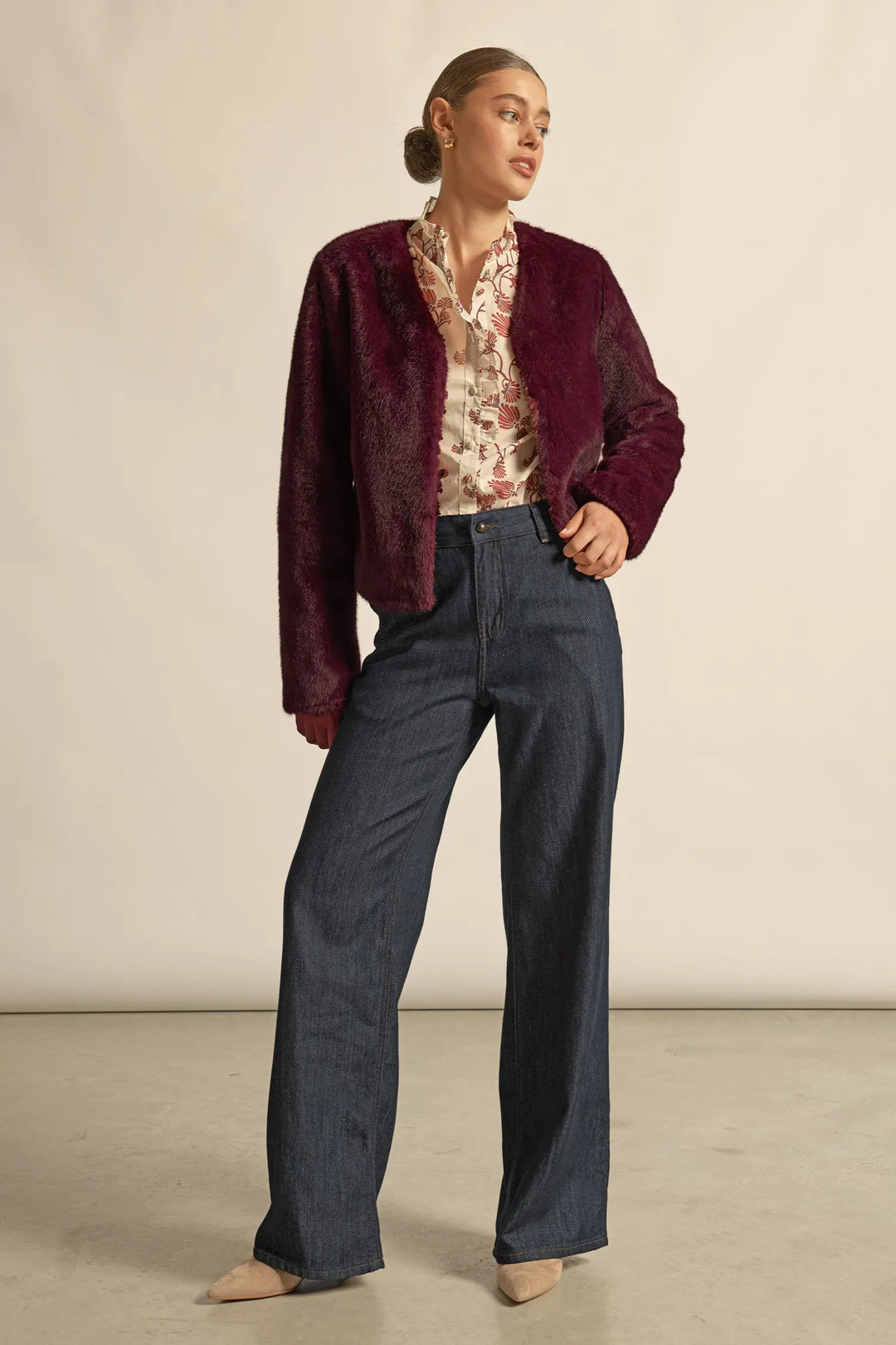 Woman wearing a burgundy jacket, patterned blouse, and dark pants against a beige background