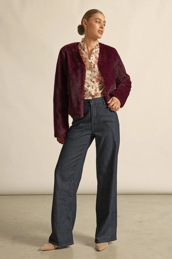 Woman wearing a burgundy jacket, patterned blouse, and dark pants against a beige background